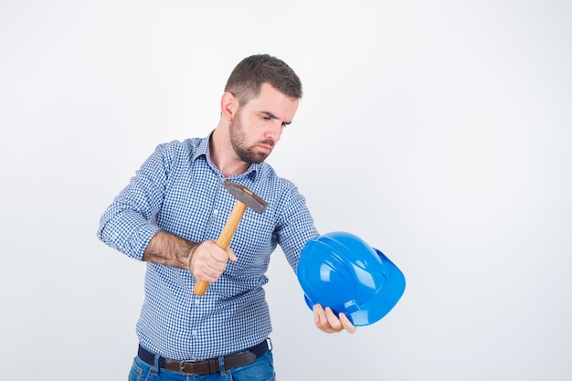 Casco llamativo masculino joven con un martillo en camisa, jeans, casco y mirando serio, vista frontal.