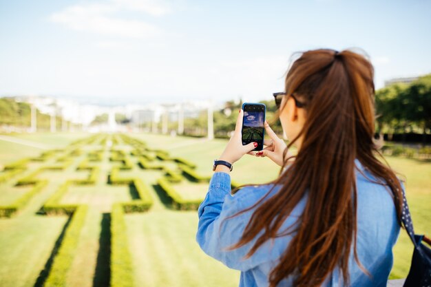 Casual hermosa joven posando para la foto del teléfono inteligente en el parque jardín de la ciudad en verano.