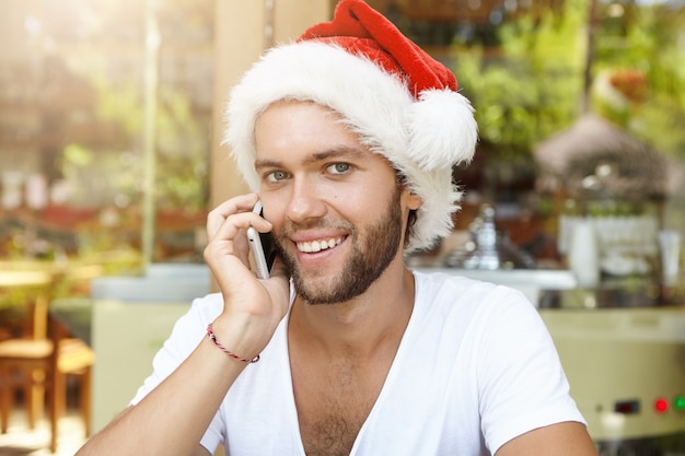 Caucásico joven alegre en sombrero de Santa Claus con conversación telefónica