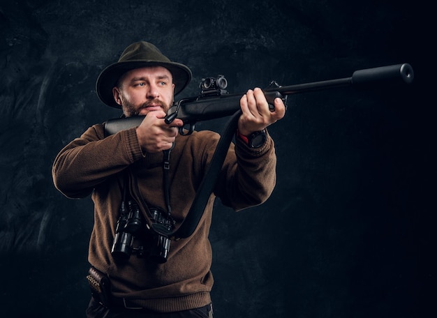 Cazador masculino sosteniendo un rifle y apuntando a su objetivo o presa. Foto de estudio contra el fondo de la pared oscura