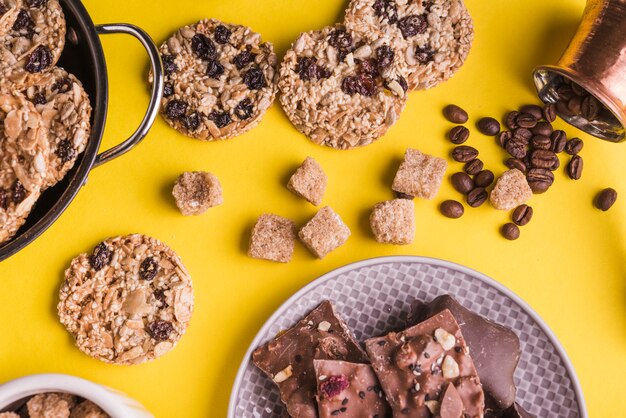 Cejas de azúcar en cubitos; galletas de chocolate; Granos de café y placa de barras de chocolate sobre fondo amarillo brillante