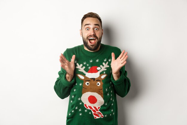 Celebración de año nuevo y concepto de vacaciones de invierno. Chico barbudo feliz y sorprendido mirando asombrado, atrapando algo, de pie en un divertido suéter de Navidad, fondo blanco.