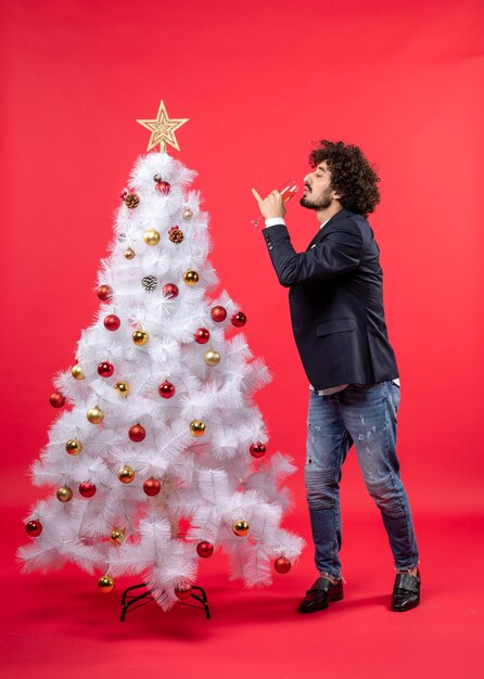 Celebración del año nuevo con un joven barbudo bebiendo vino y de pie cerca del árbol de Navidad blanco decorado
