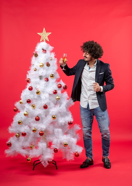 Celebración del año nuevo con un joven barbudo sosteniendo una copa de vino y de pie cerca del árbol de Navidad blanco decorado