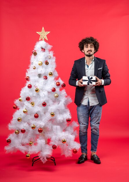 Celebración del año nuevo con un joven serio sosteniendo un regalo cerca del árbol de Navidad blanco decorado en imágenes rojas