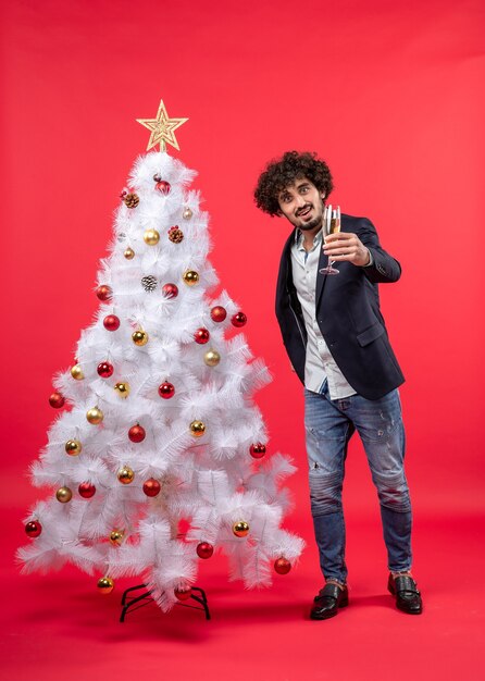 Celebración del año nuevo con joven sosteniendo una copa de vino cerca del árbol de Navidad blanco decorado en imágenes rojas