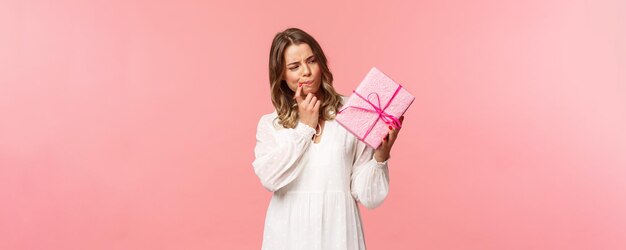 Celebración de días festivos y concepto de mujer Retrato de una chica rubia curiosa y pensativa pensando en lo que hay dentro de la caja de regalo, reflexionando sobre decir hmm como haciendo una suposición de fondo rosa