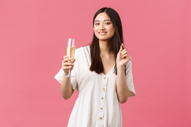 Celebración, fiestas y concepto de diversión. Elegante mujer joven y bonita asistir al evento, bebiendo champán y sonriendo con alegría, disfrutando de la celebración, de pie con un vestido blanco sobre fondo rosa.