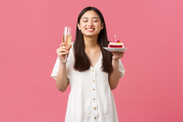 Celebración, fiestas y concepto de diversión. Hermosa mujer soñadora celebrando su cumpleaños con copa de champán y b-day cake, sonriendo y pidiendo deseos sobre fondo rosa.