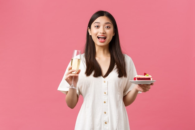 Celebración, fiestas y concepto de diversión. Mujer hermosa soñadora celebrando su cumpleaños con copa de champán y b-day cake, mirando sorprendido y emocionado, de pie con fondo rosa.