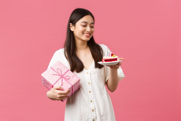 Celebración, fiestas y concepto de diversión. Soñadora feliz cumpleañera en vestido blanco, sonriendo y mirando a otro lado como recibiendo regalo, comiendo pastel de cumpleaños, fondo rosa