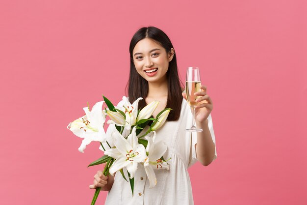 Celebración, fiestas y concepto de diversión. Sonriente mujer asiática bastante glamour en vestido con ramo de lirios blancos, levantando una copa de champán para hacer un brindis, bebida para la cumpleañera