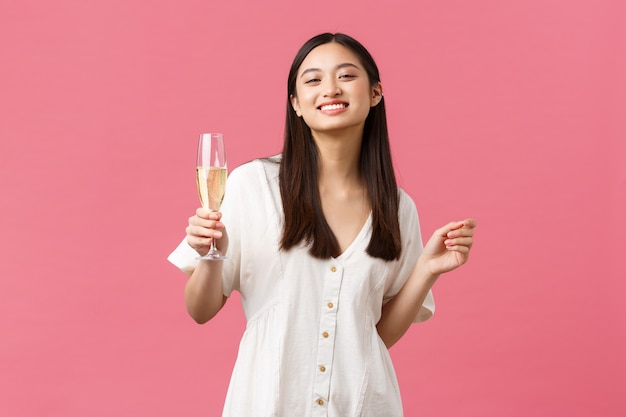 Celebración, fiestas y concepto de diversión. Sonriente niña feliz cumpleaños en vestido blanco, disfrutando celebrando con amigos, sosteniendo una copa de champán sobre fondo rosa.