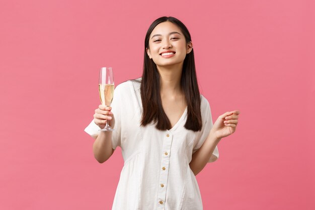 Celebración, fiestas y concepto de diversión. Sonriente niña feliz cumpleaños en vestido blanco, disfrutando celebrando con amigos, sosteniendo una copa de champán sobre fondo rosa.