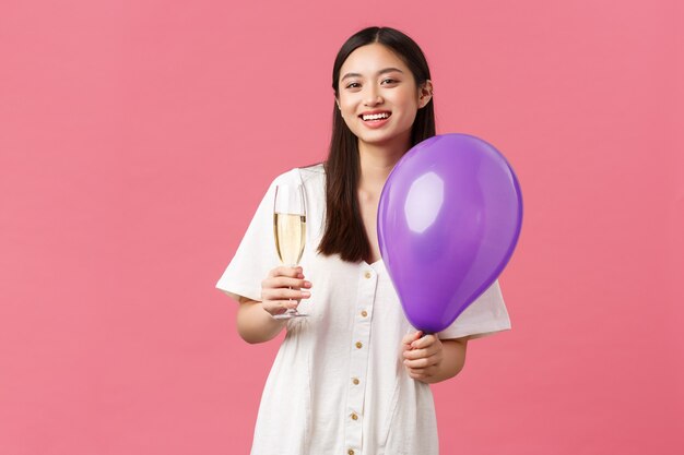 Celebración, fiestas y concepto de diversión. Tierna mujer bastante asiática en vestido blanco, sosteniendo un globo y una copa de champán, haciendo tostadas durante el evento, disfrutando del momento, fondo rosa.