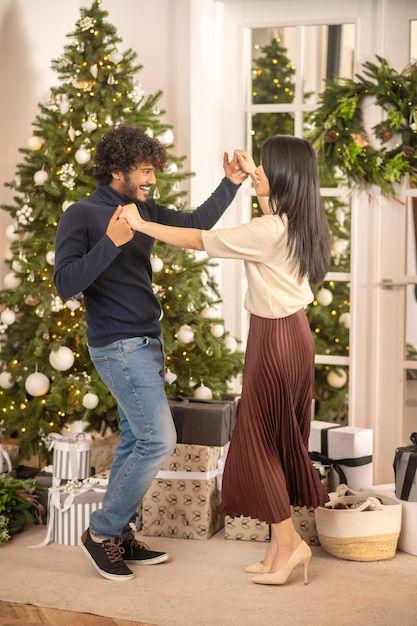 Celebracion. Joven indio sonriente barbudo y mujer bonita asiática con tacones altos mirándose bailando tocando la mano cerca del árbol de navidad y regalos
