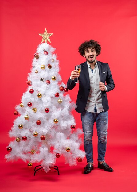 Celebración de Navidad con barbudo joven feliz con vino de pie cerca del árbol de Navidad