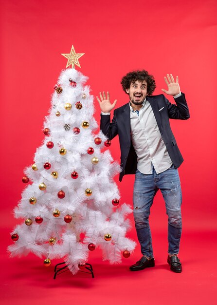 Celebración de Navidad con feliz emocionado joven haciendo reacciones divertidas y de pie cerca del árbol de Navidad