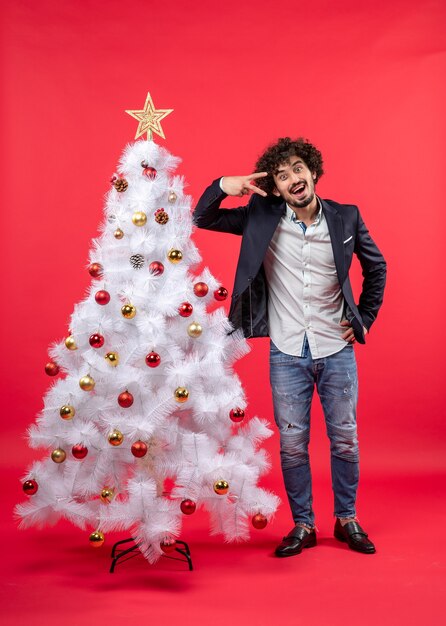 Celebración de Navidad con feliz emocionado joven mostrando dos y de pie cerca del árbol de Navidad