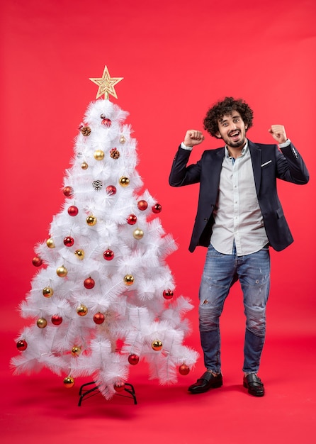 Celebración de Navidad con feliz emocionado joven orgulloso de sí mismo y de pie cerca del árbol de Navidad