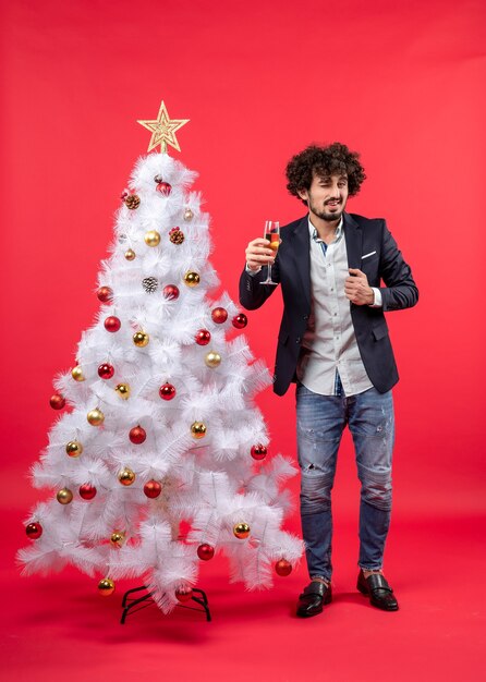 Celebración de Navidad con joven barbudo con vino diciendo algo de pie cerca del árbol de Navidad en rojo