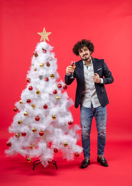 Celebración de Navidad con un joven barbudo con winelooking a la cámara y de pie cerca del árbol de Navidad en rojo