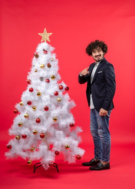 Celebración de Navidad con joven gracioso emocionado feliz apuntando algo de pie cerca del árbol de Navidad