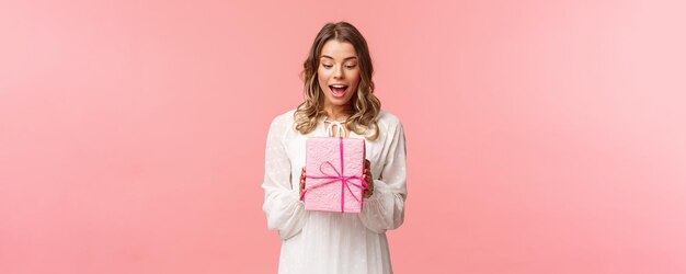 Celebración navideña y concepto de mujer Retrato de una encantadora joven rubia sorprendida recibe un regalo sorpresa sosteniendo un regalo en una caja rosa míralo divertido curioso qué hay dentro del fondo del estudio