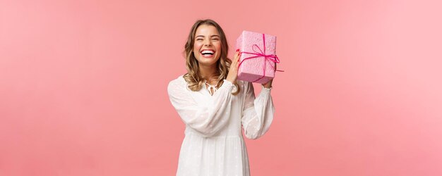 Celebración navideña y concepto de mujer Retrato de feliz carismática rubia sacudiendo caja de regalo preguntándose qué hay dentro mientras celebra cumpleaños recibe regalos de cumpleaños fondo rosa