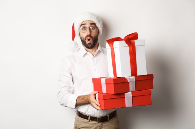 Celebración y vacaciones de invierno. Hombre emocionado sosteniendo regalos de Navidad y mirando sorprendido, vistiendo gorro de Papá Noel, de pie