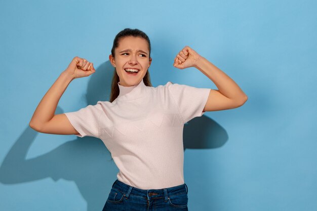 Celebrando como ganador. Retrato de la muchacha adolescente caucásica sobre fondo azul. Preciosa modelo en ropa casual. Concepto de emociones humanas, expresión facial, ventas, publicidad. Copyspace. Se ve lindo, asombrado.