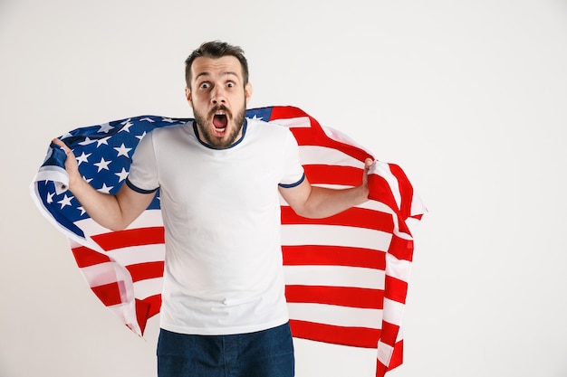 Celebrando un día de la Independencia. Estrellas y rayas. Hombre joven con la bandera de los Estados Unidos de América aislado en la pared blanca del estudio. Parece loco feliz y orgulloso como un patriota de su país.