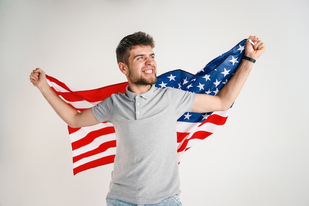 Foto gratuita celebrando un día de la independencia. estrellas y rayas. hombre joven con la bandera de los estados unidos de américa aislado en la pared blanca del estudio. parece loco feliz y orgulloso como un patriota de su país.