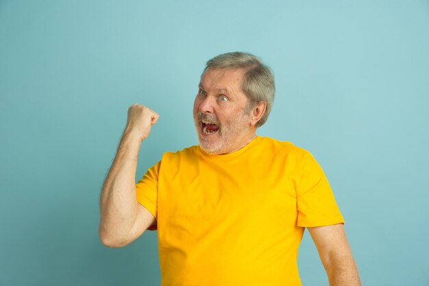 Celebrando ganar, deporte. Retrato de hombre caucásico aislado sobre fondo azul de estudio. Hermoso modelo masculino en camisa amarilla posando.