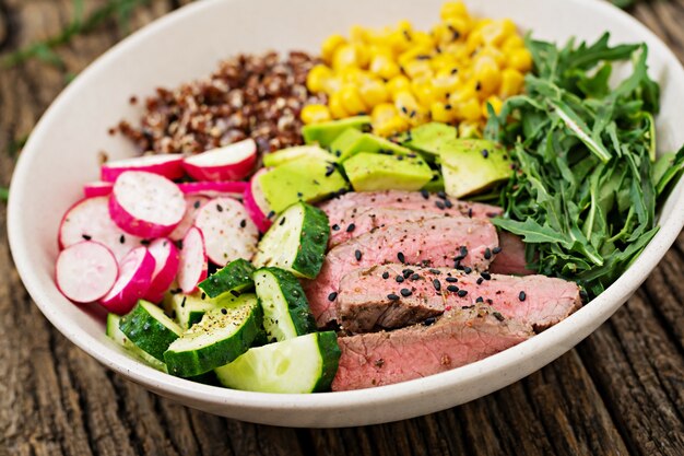 Cena Saludable. Buda tazón almuerzo con filete de ternera a la parrilla y quinua, maíz, aguacate, pepino y rúcula en mesa de madera. Ensalada de carne