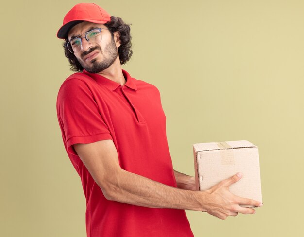 Con el ceño fruncido joven repartidor caucásico en uniforme rojo y gorra con gafas de pie en la vista de perfil sosteniendo cardbox aislado en la pared verde oliva