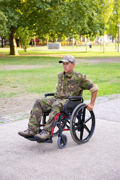 Centrado militar discapacitado en silla de ruedas con uniforme de camuflaje, bajando por la acera en el parque de la ciudad. Veterano de guerra o concepto de discapacidad