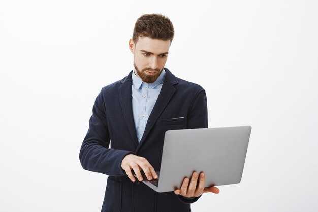 Centrándose en los negocios. Retrato de inteligente y ambicioso, concentrado, apuesto joven empresario con barba y ojos azules, sosteniendo una computadora portátil en la mano, revisando el horario con una mirada determinada