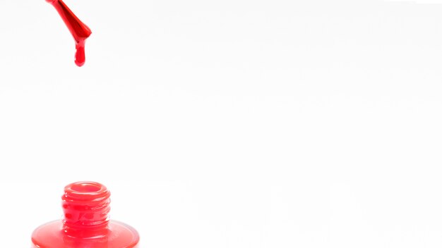 Cepillo de esmalte de uñas rojo con una gota cayendo en la botella sobre fondo blanco