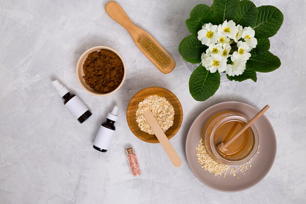 Cepillo de madera; miel; avena; sal de roca del Himalaya; Botella de aceite esencial con jarrón de flores de primula sobre fondo de hormigón