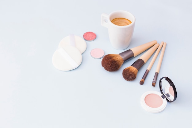 Cepillo de maquillaje con esponja; sombra de ojos y colorete con taza de café sobre fondo blanco