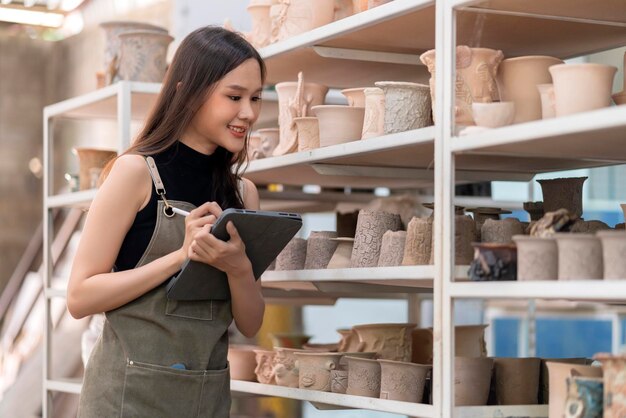 La ceramista asiática con la mano del delantal arregla su producto de escultura de arcilla en el estante esperando hornear en el horno mujer asiática dueña de un pequeño taller de cerámica sonriendo orgullosa de presentar su nueva colección