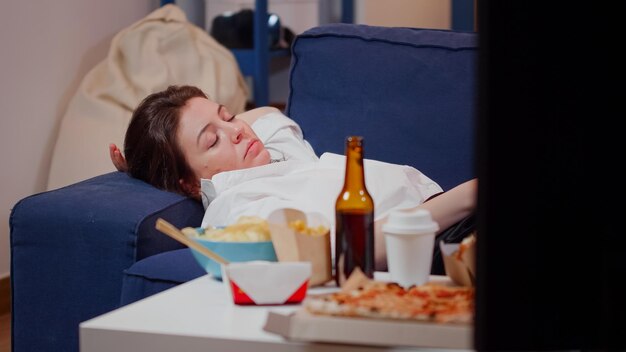 Cerca de adulto cansado tendido en el sofá con el control remoto del televisor en la mano en casa. Mujer dormida en el sofá en la sala de estar mientras ve la televisión después de comer comida rápida para llevar