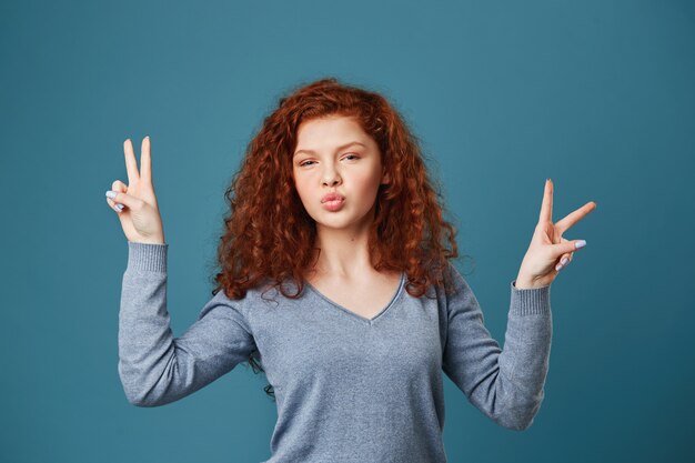 Cerca de alegre joven con pelo rojo ondulado y pecas con gesto de paz en ambas manos, haciendo labios de pato posando para la foto en la fiesta.