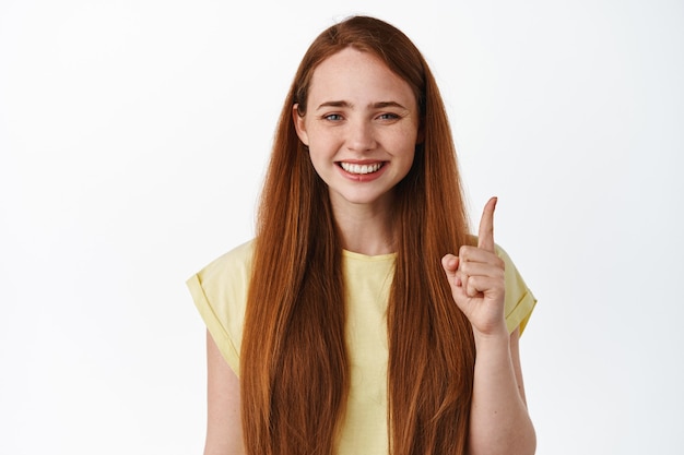 Cerca de alegre joven con pelo rojo y sonrisa blanca, apuntando con el dedo hacia arriba de pie en camiseta en blanco.