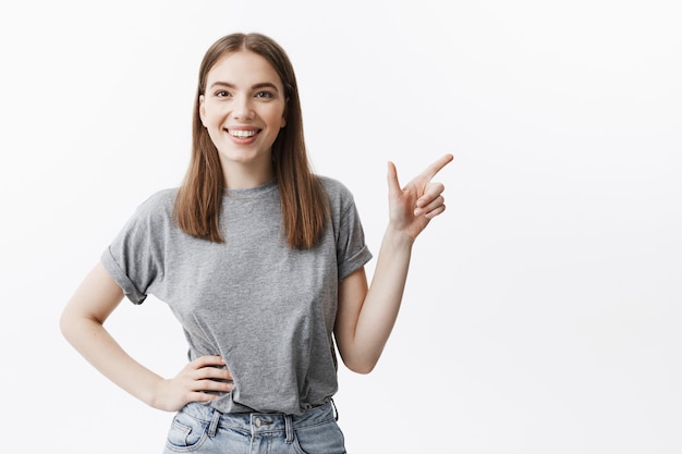 Cerca de apuesto chica alegre estudiante europeo con cabello oscuro en ropa casual sonriendo con expresión de cara alegre, apuntando a un lado con el dedo en el espacio libre