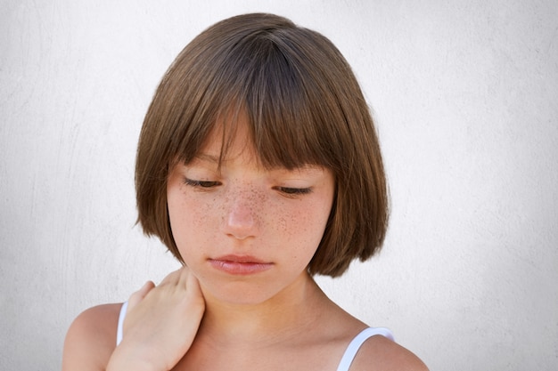 Cerca de atractivo niño pequeño con pecas y cabello corto oscuro manteniendo su mano en el cuello, mirando seriamente hacia abajo, con expresión pensativa mientras posa en la pared blanca de concerete