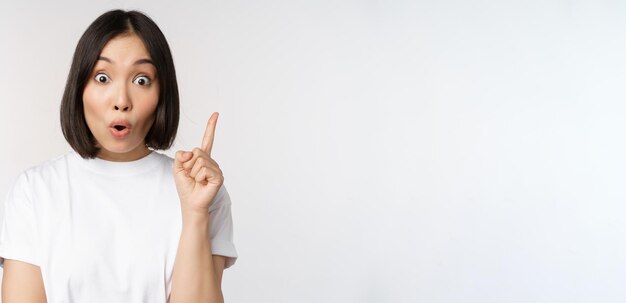 Cerca de una chica asiática creativa levantando el dedo sugiriendo que algo tiene una idea apuntando hacia el cartel de eureka de pie sobre fondo blanco