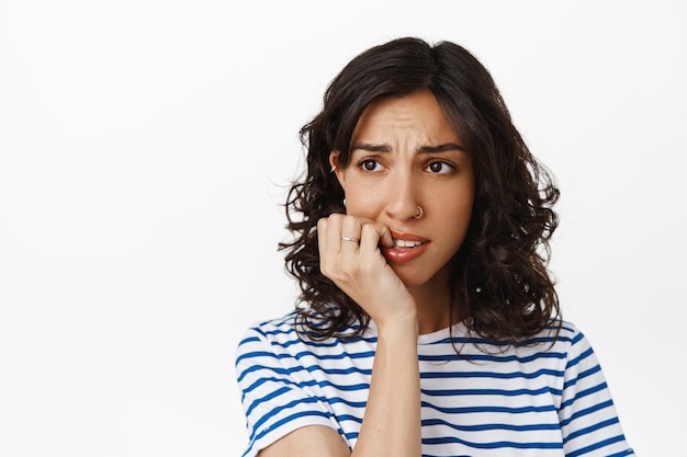 Cerca de una chica morena ansiosa, mordiéndose las uñas y mirando hacia otro lado con cara de alarma y nerviosismo, sintiendo miedo a las consecuencias, teniendo miedo, de pie en blanco