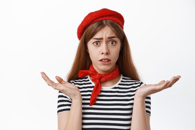 Cerca de confundida chica francesa con boina y camiseta a rayas, encogiéndose de hombros con cara de perplejidad, no puedo entender algo, de pie sobre fondo blanco.